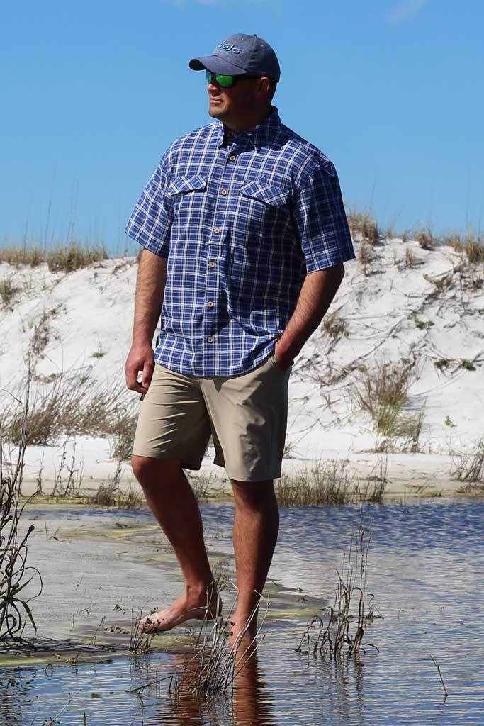 Man standing in water wearing a blue plaid shirt and beige shorts with a clear sky background.