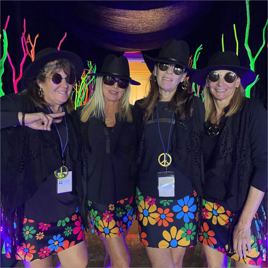 Four women wearing sunglasses and black hats with colorful flower-patterned skirts in front of a vibrant, abstract background.