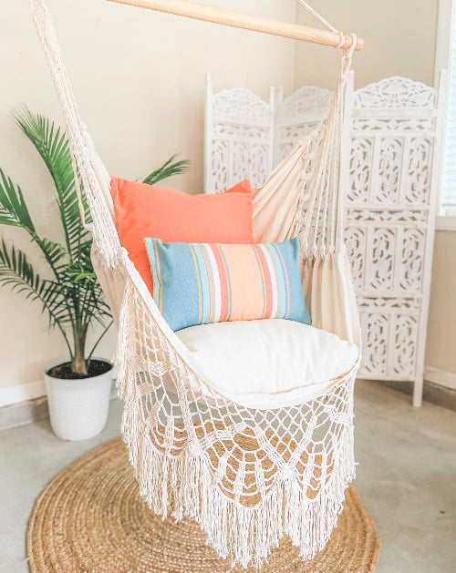 White hammock chair with colorful pillows in a room with a plant and decorative lattice wall.