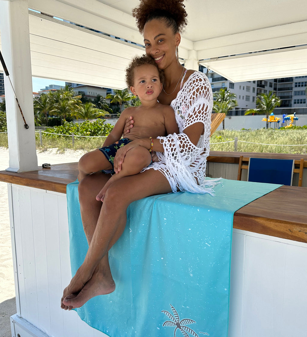 Woman and child sitting on a turquoise towel with a beach view