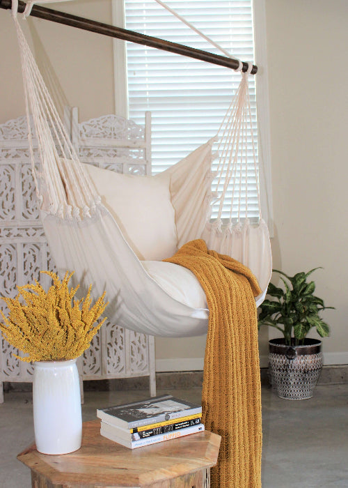 White hammock chair with a yellow blanket in a room with a wooden table, books, and plants.