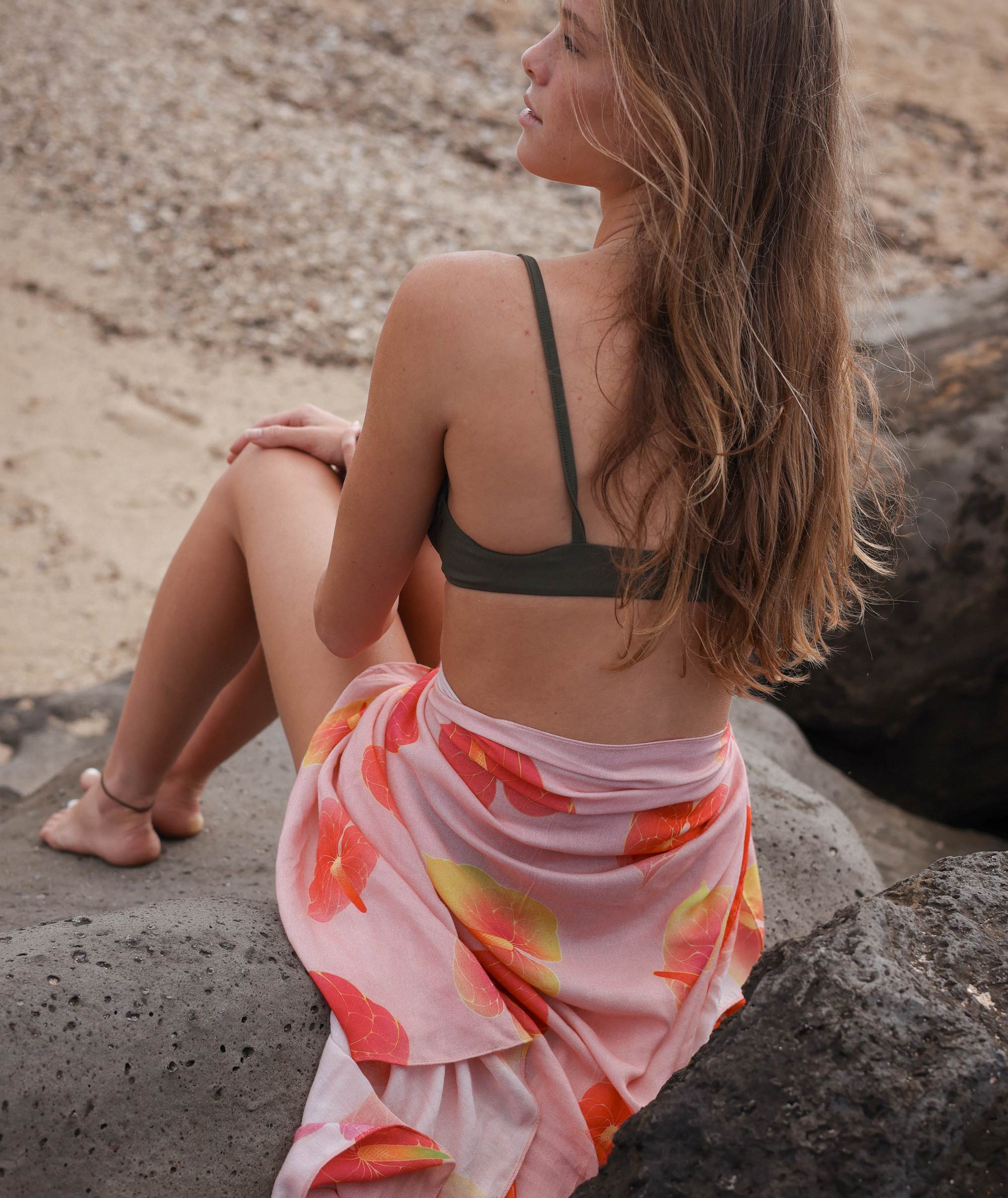 Woman sitting on rocks wearing a black bikini top and colorful sarong.