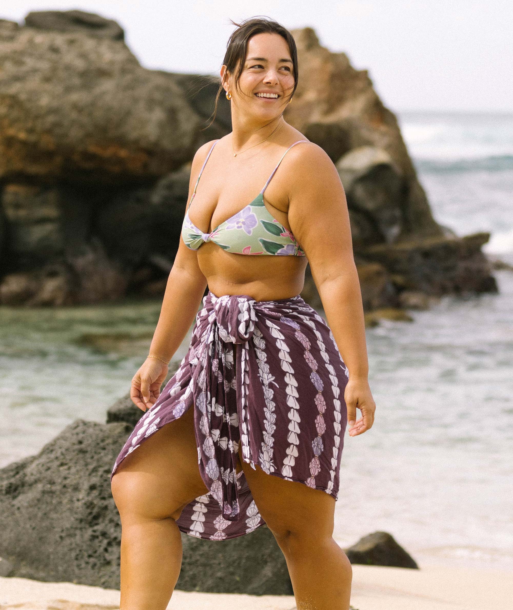 Woman in a floral bikini top and purple patterned sarong standing on a beach with rocks and water in the background.