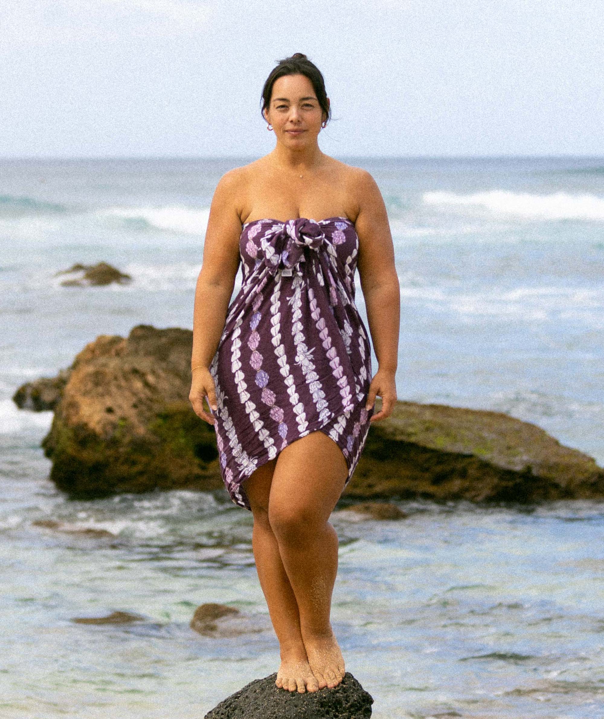 Woman in a purple patterned dress standing on a rock by the ocean.