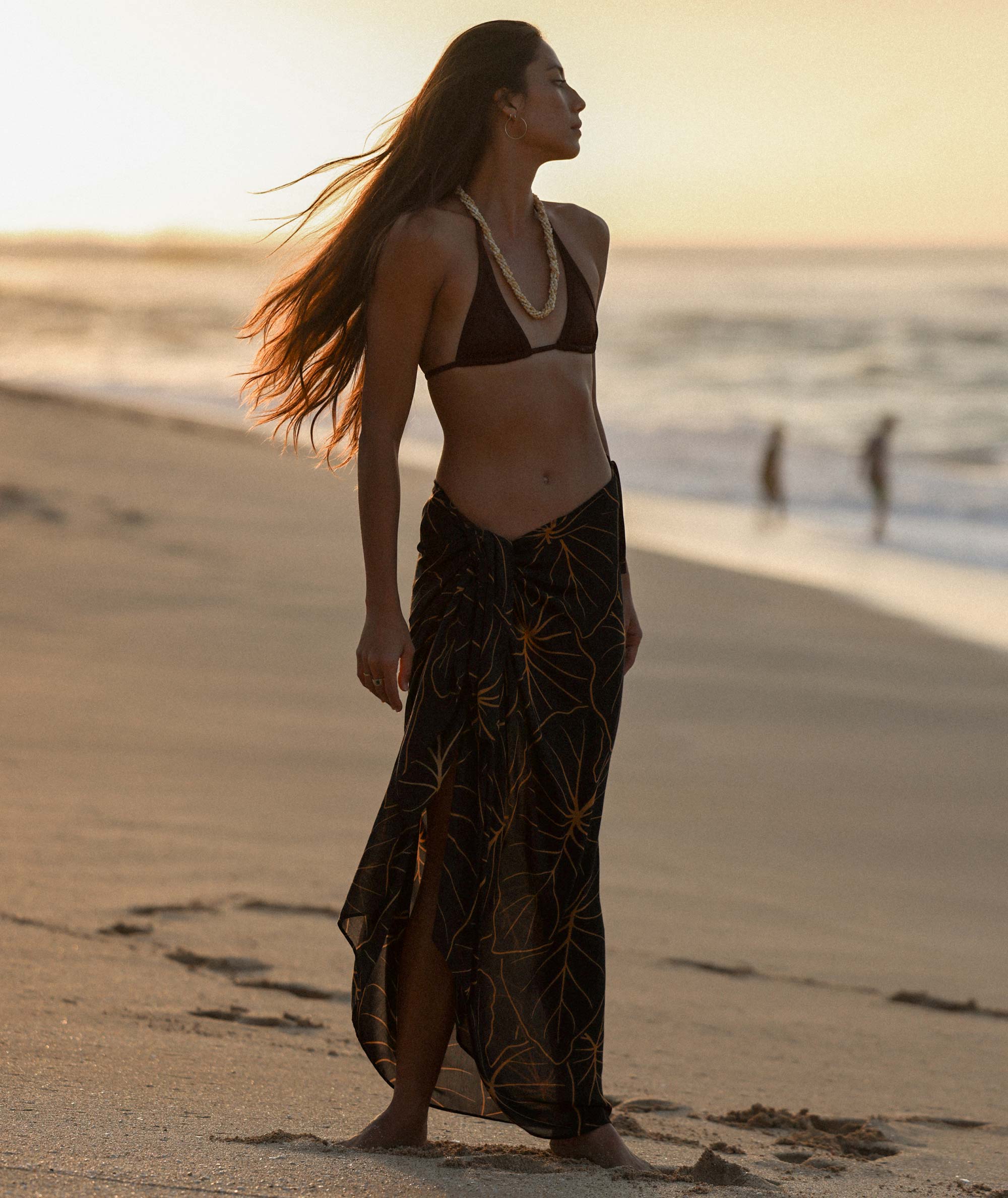 Woman walking on a beach at sunset wearing a brown bikini top and black patterned sarong.