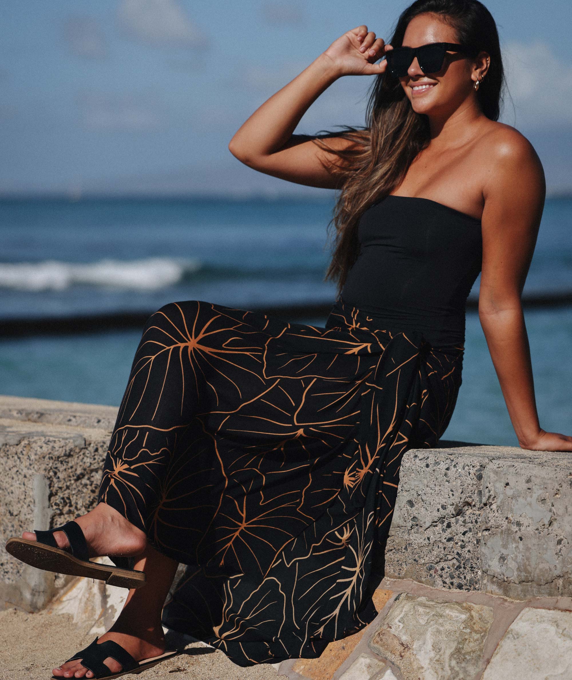 Woman in a black dress with gold leaf pattern sitting by the ocean.