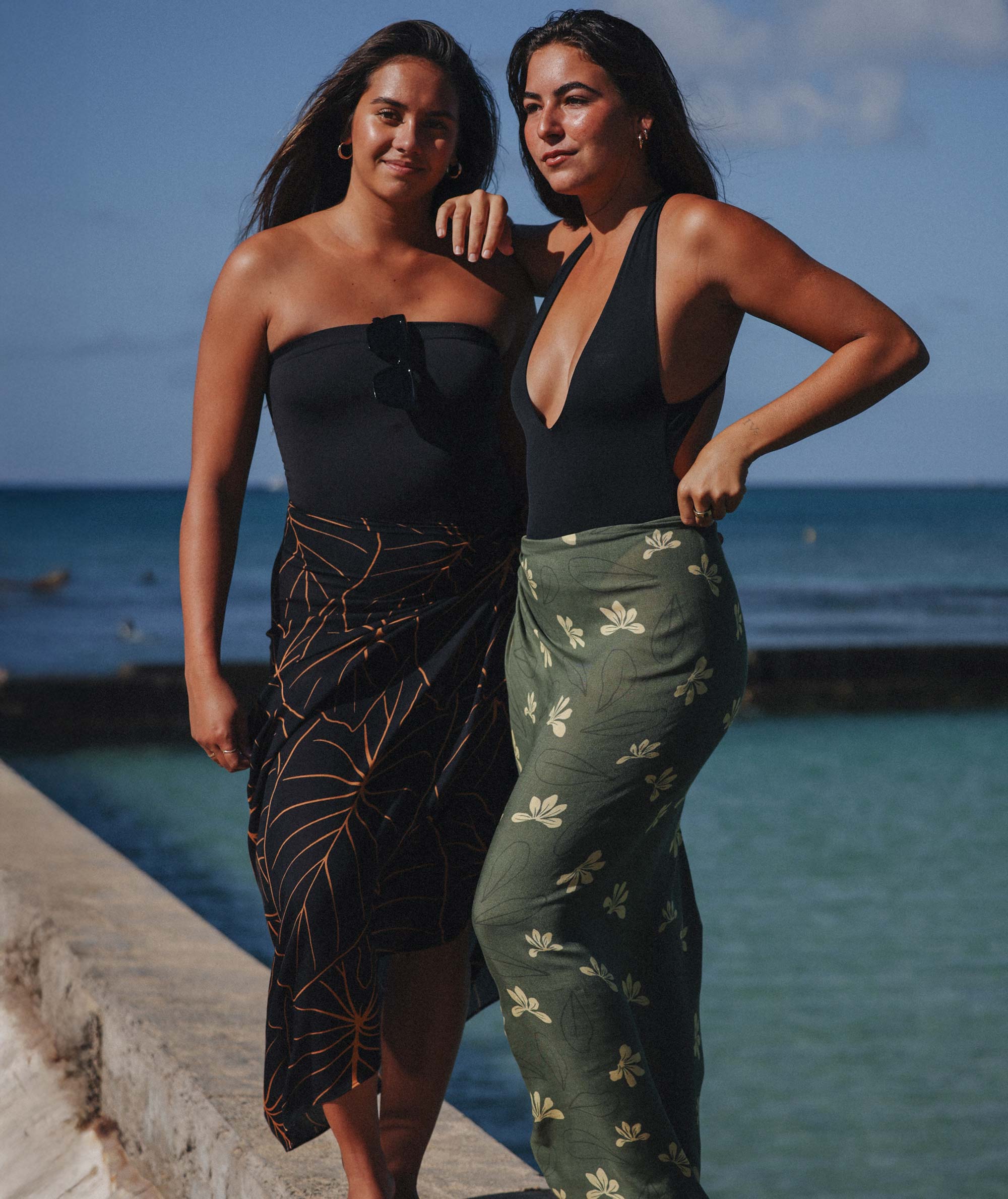 Two women standing by the ocean wearing black and green dresses.