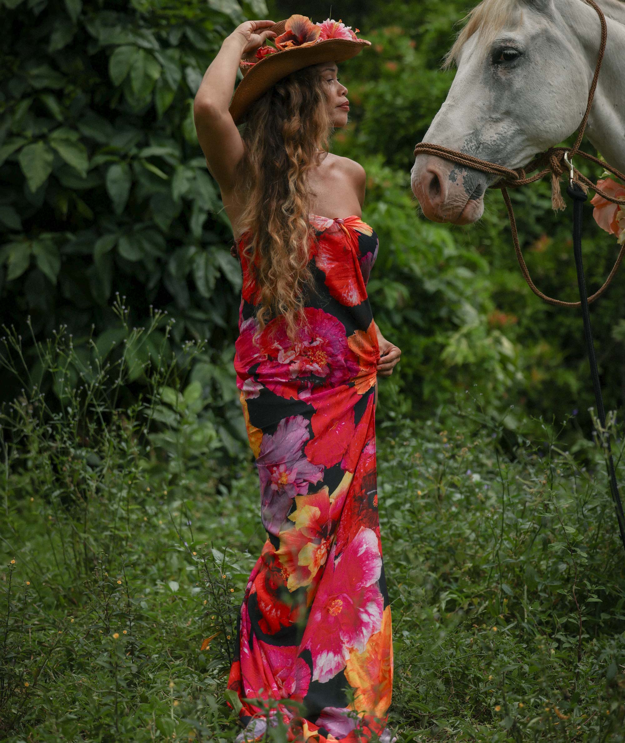 Woman in a colorful floral dress standing next to a white horse in a grassy area.