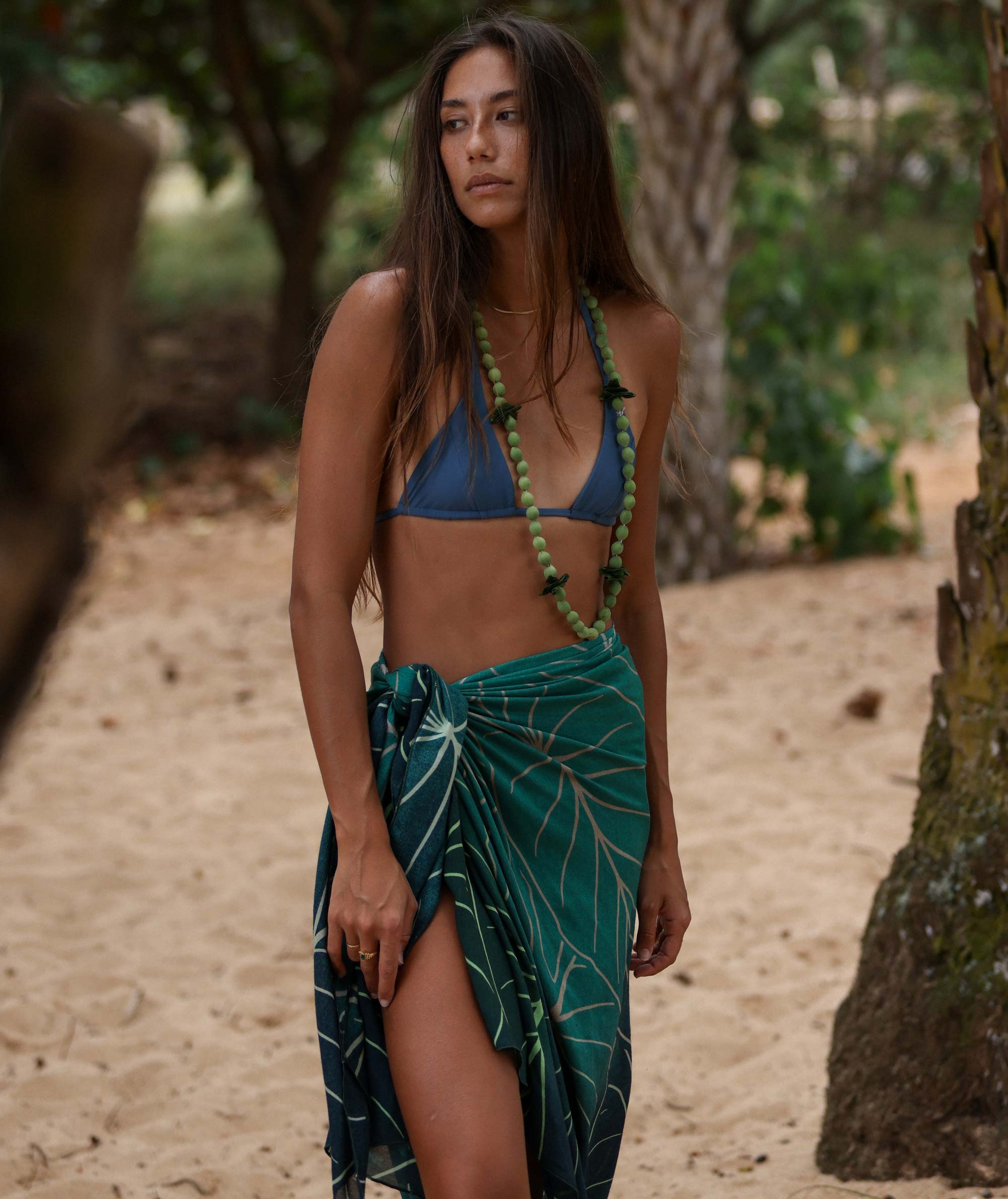 Woman in a blue bikini top and green leaf-patterned sarong standing on a sandy path with trees in the background.