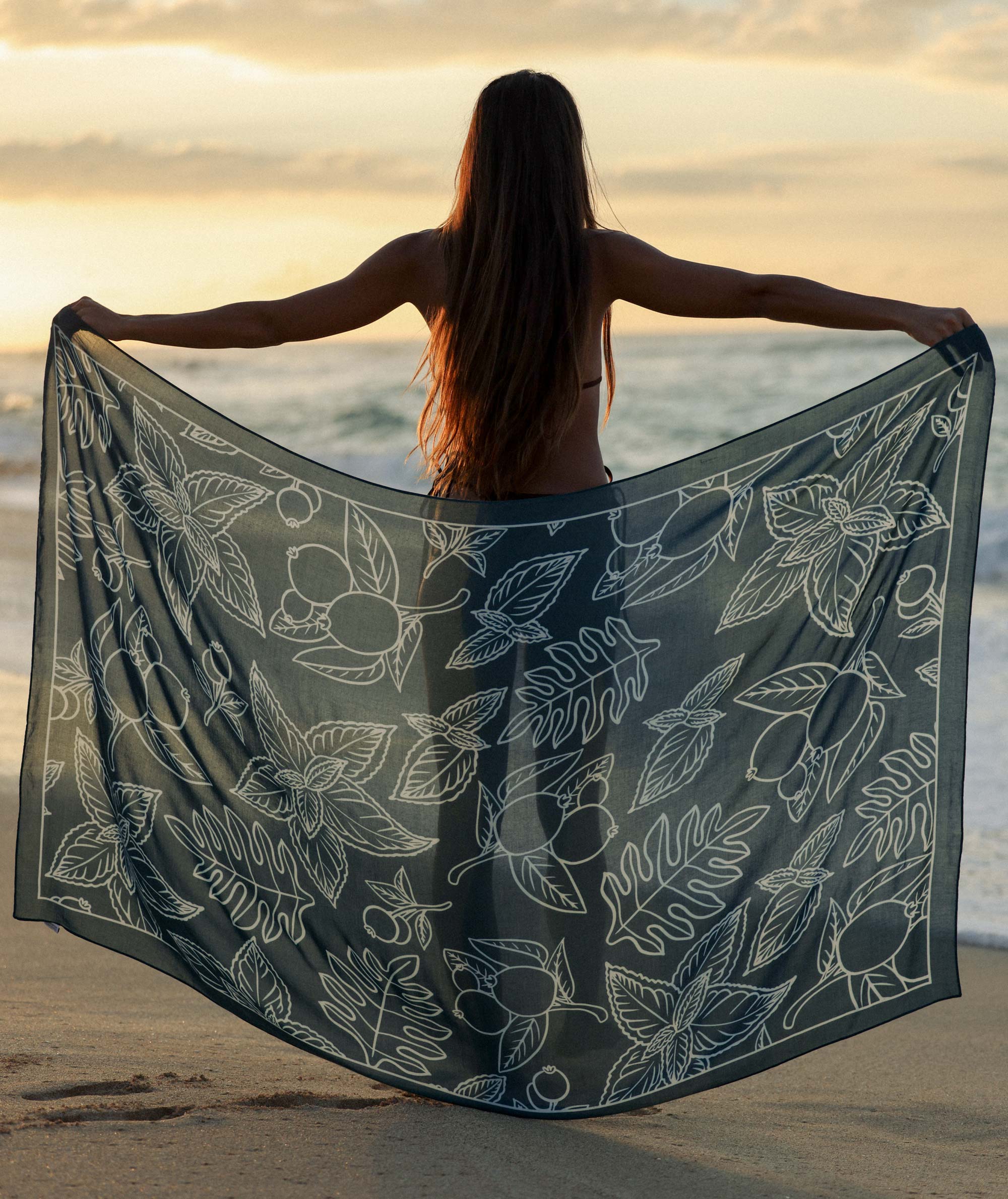 Woman on a beach holding a floral-patterned towel with a sunset in the background