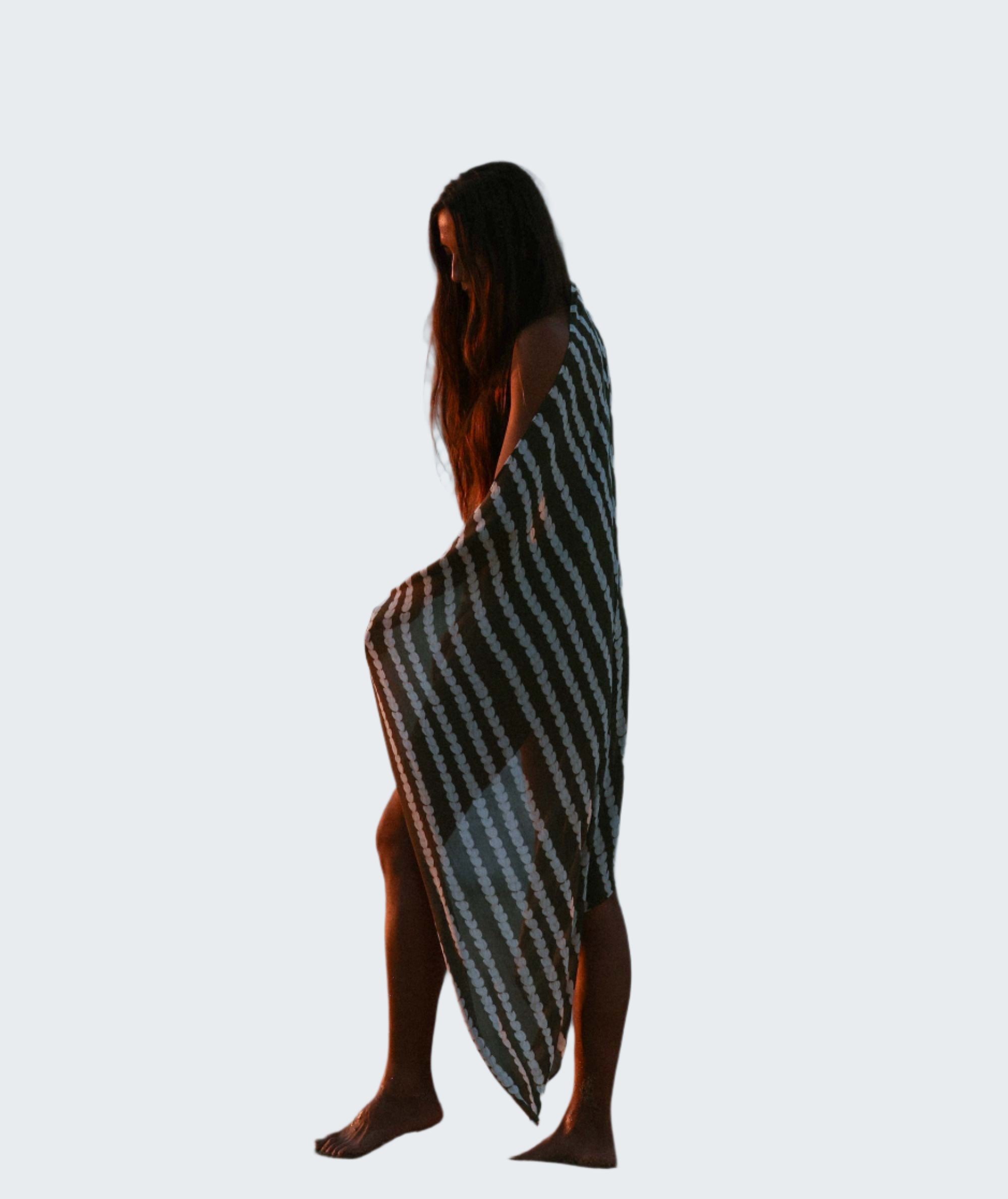 Woman standing on a beach with a striped towel draped over her, facing the ocean.