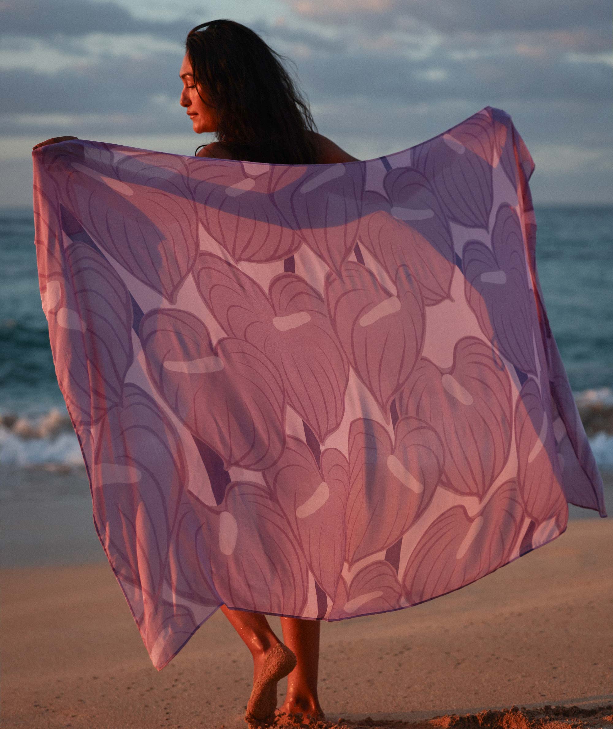 Woman holding a purple floral patterned scarf on a beach at sunset