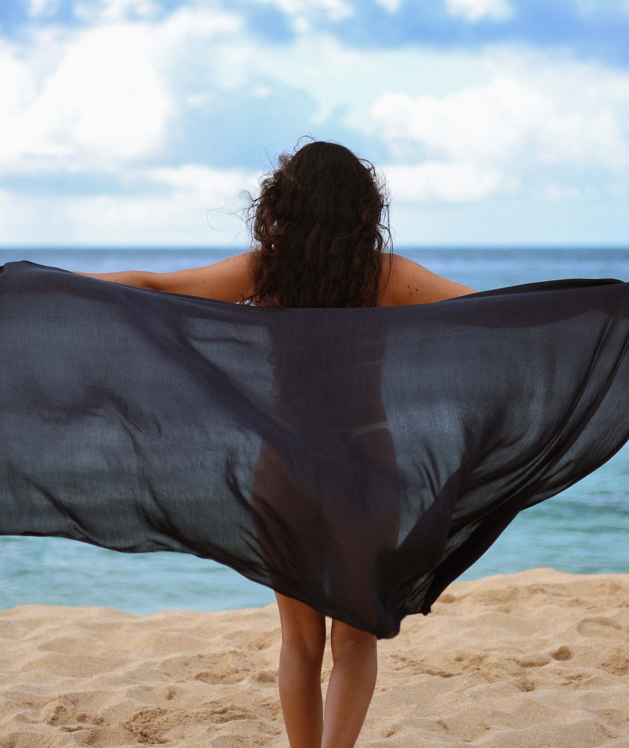 Person standing on a beach with a black fabric flowing in the wind, looking out at the ocean.