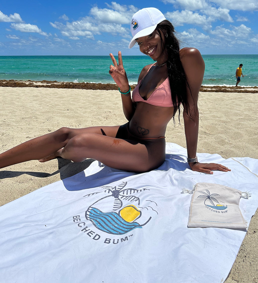 Woman in a bikini and cap sitting on a towel with 'Beached Bum' branding at the beach.