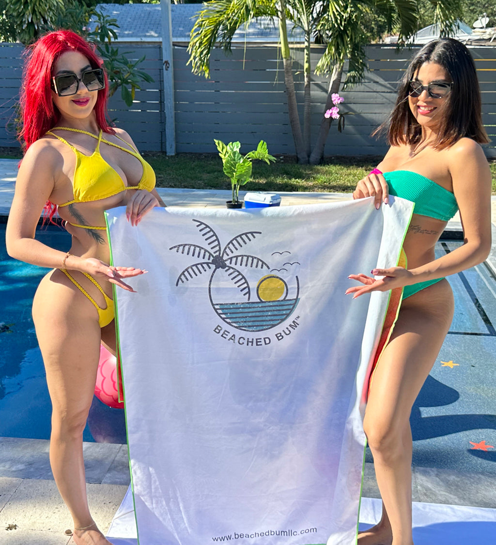 Two women in bikinis holding a towel with a palm tree and sun design by a pool.