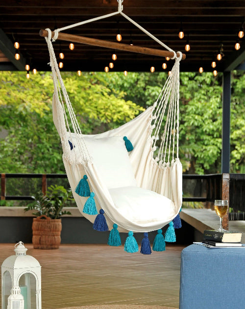 White hammock chair with blue tassels hanging in an outdoor setting with greenery and a wooden floor.