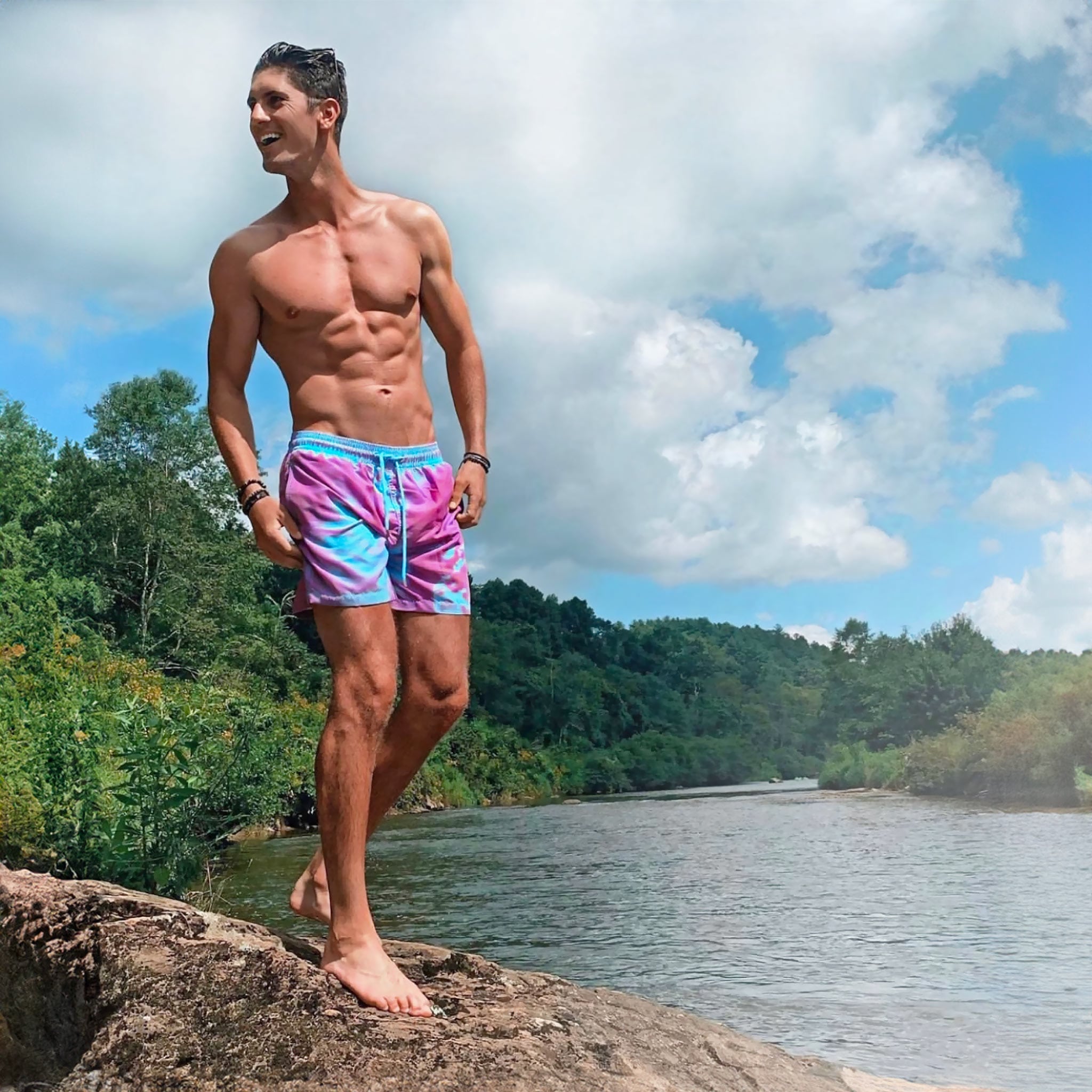 Man in colorful swim shorts standing on a rock by a lake with trees in the background