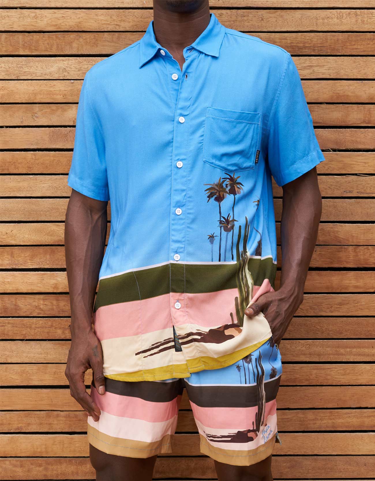 Person wearing a blue shirt with palm tree design and colorful shorts against a wooden background