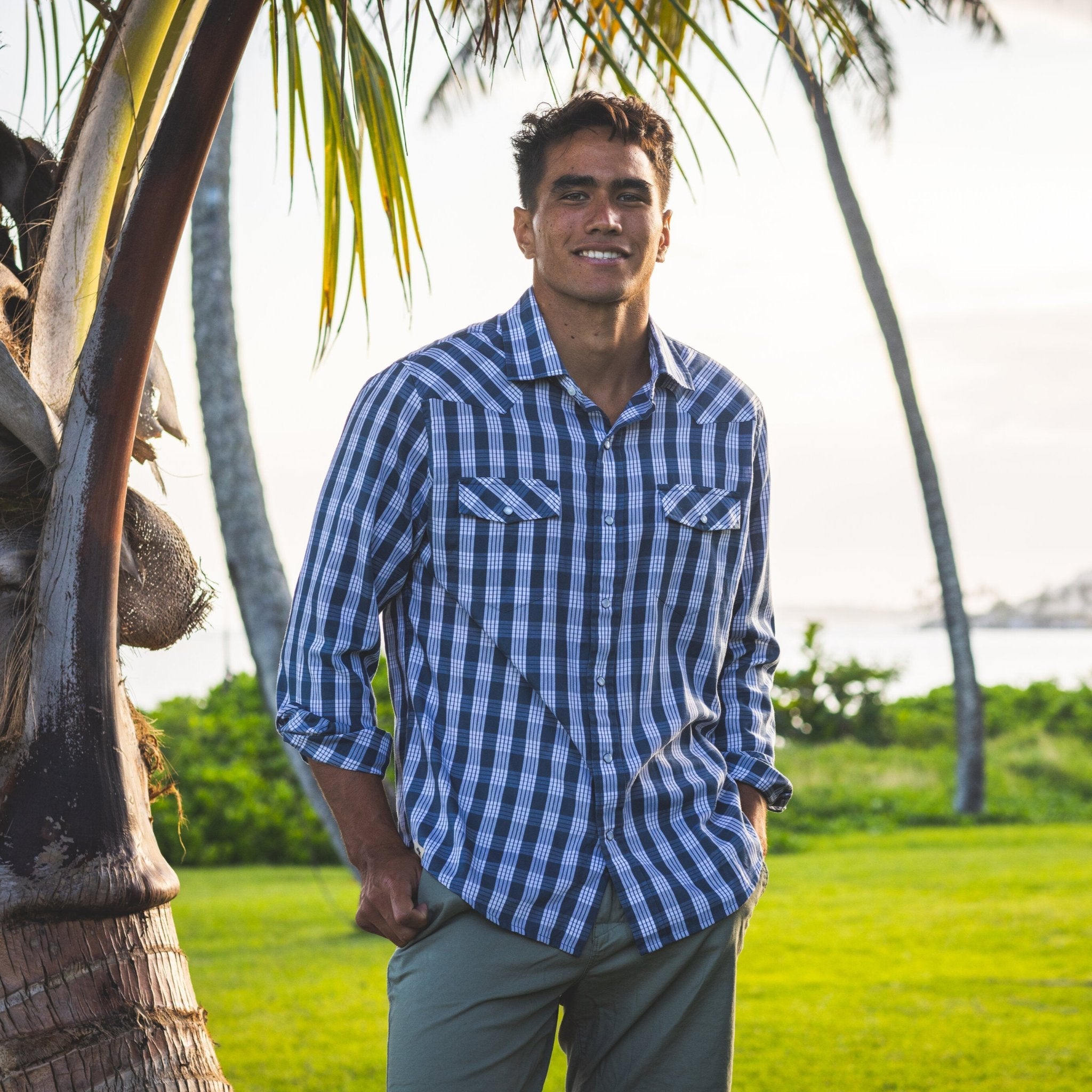 Man in a blue checkered shirt standing in a grassy area with palm trees.