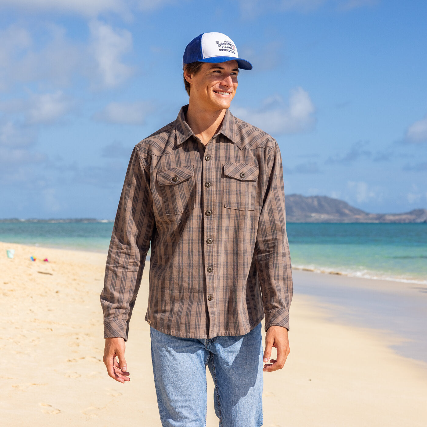 Person wearing a plaid shirt and cap on a beach with clear blue sky and ocean.