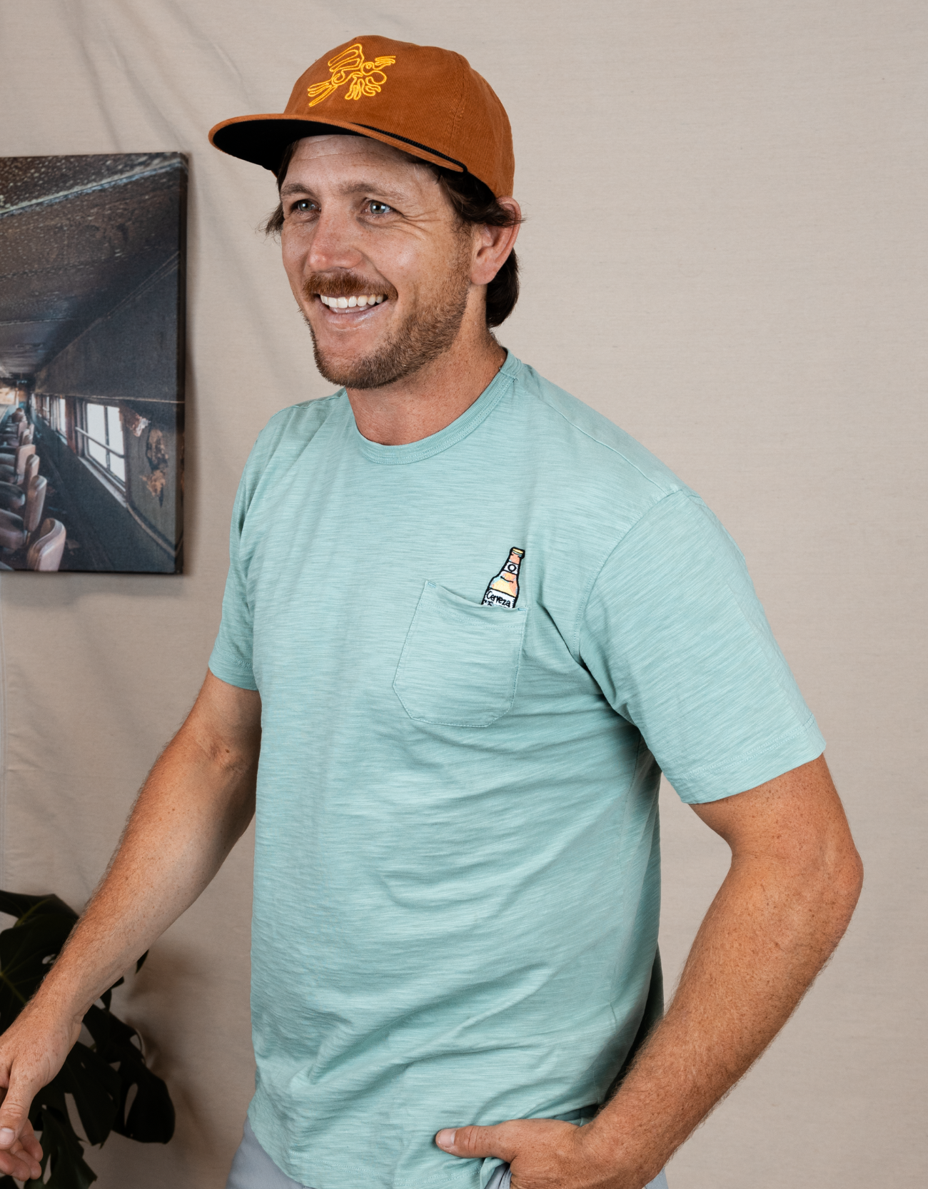 Man wearing a light blue t-shirt with a logo and a brown cap with a yellow logo, standing against a neutral background.