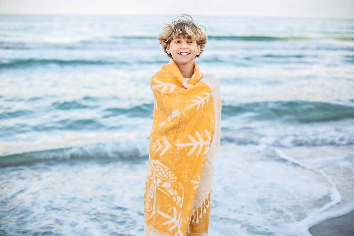 Child wrapped in a yellow towel with white patterns standing on a beach.