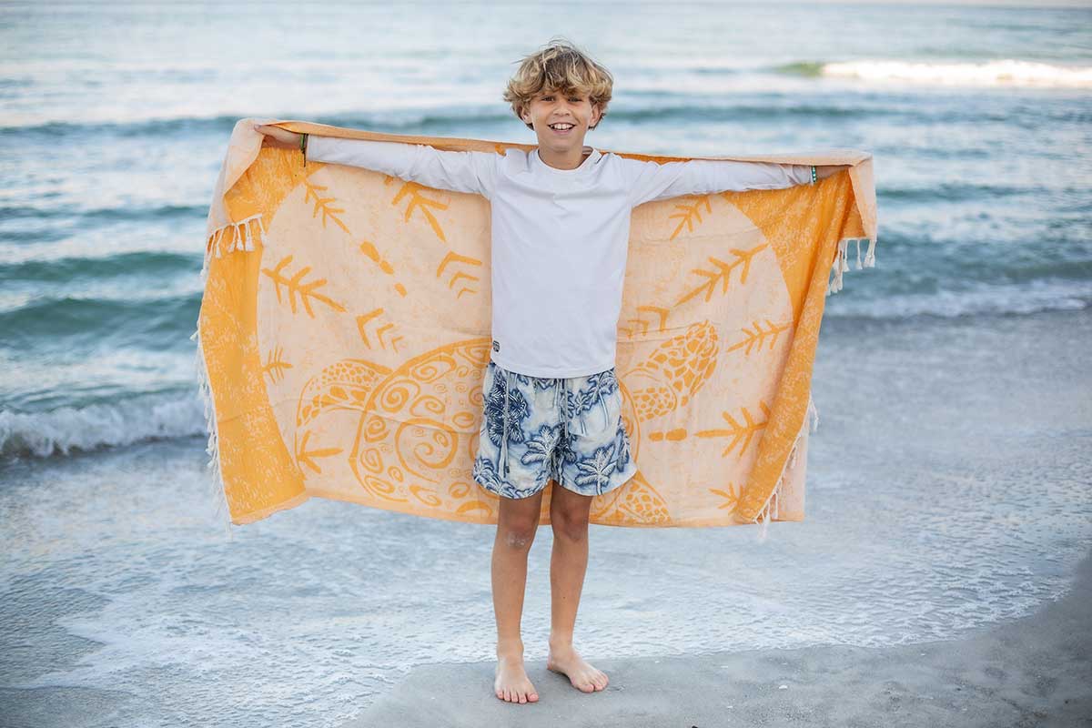 Child holding a large orange towel with patterns on a beach