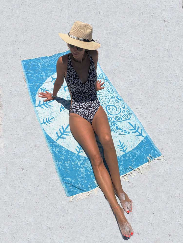 Woman in a swimsuit and hat lying on a blue beach towel with a white pattern.