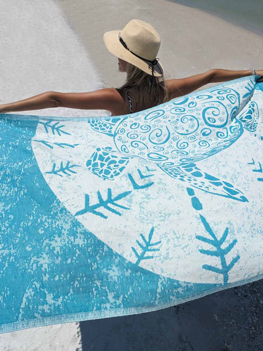 Person lying on a beach towel with a turtle design, wearing a hat.