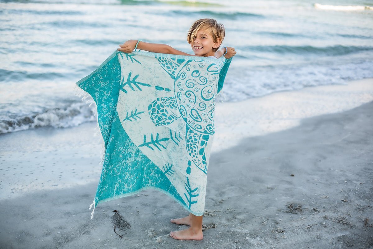 Child holding a teal and white towel with a turtle design on a beach.