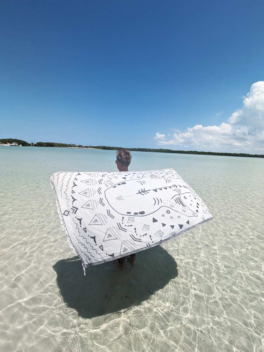 Person holding a white towel with black patterns in shallow water under a clear blue sky.