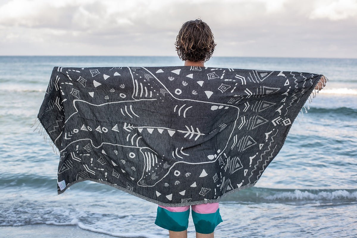 Person standing on a beach with a dark gray towel featuring white geometric patterns draped over their shoulders.