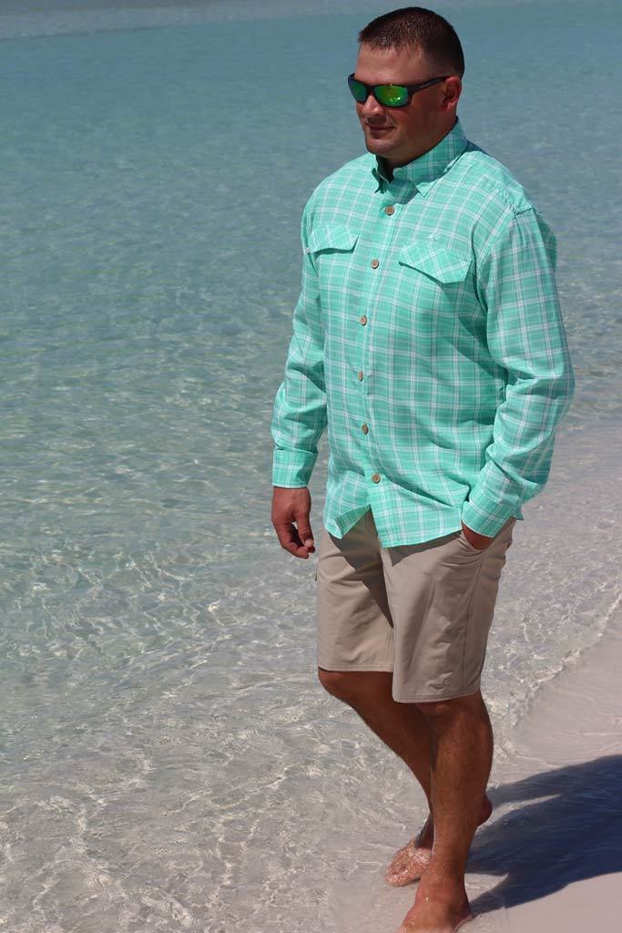 Man wearing a light green checkered shirt and beige shorts standing on a beach.