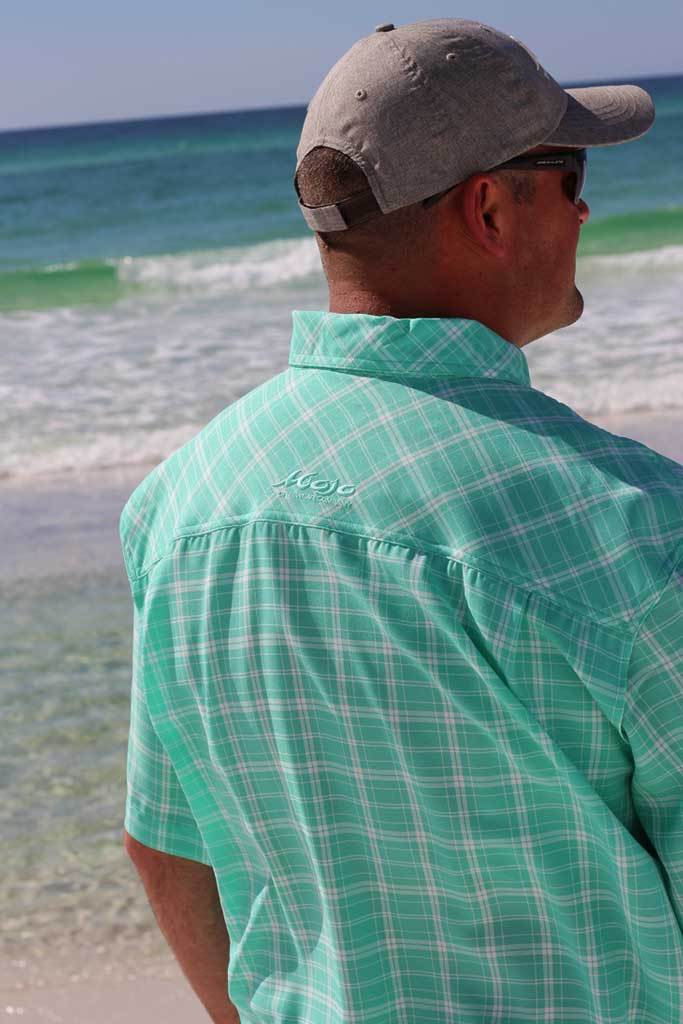 Man wearing a green plaid shirt and gray cap standing on a beach.