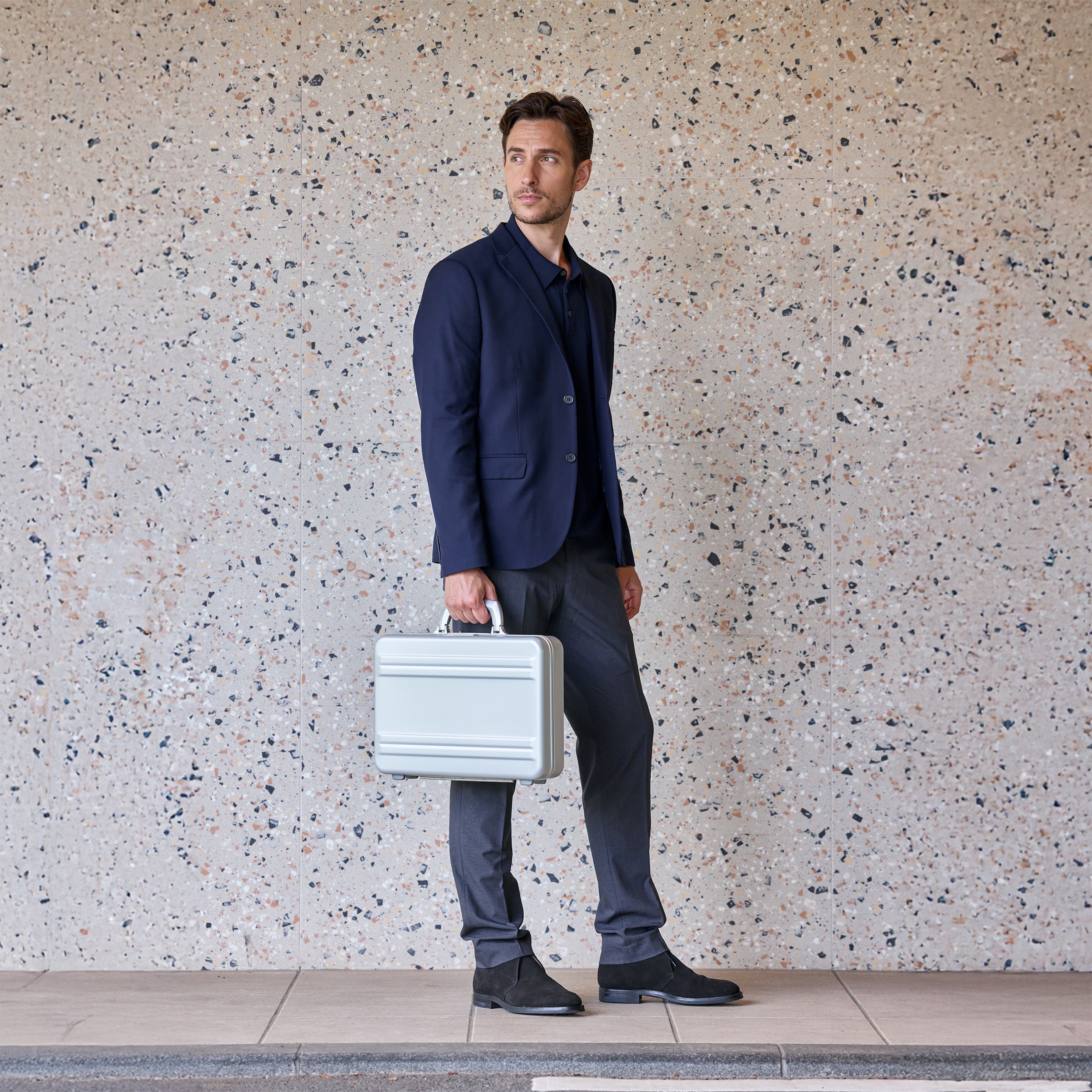 Man in a navy blazer holding a silver briefcase against a textured wall.