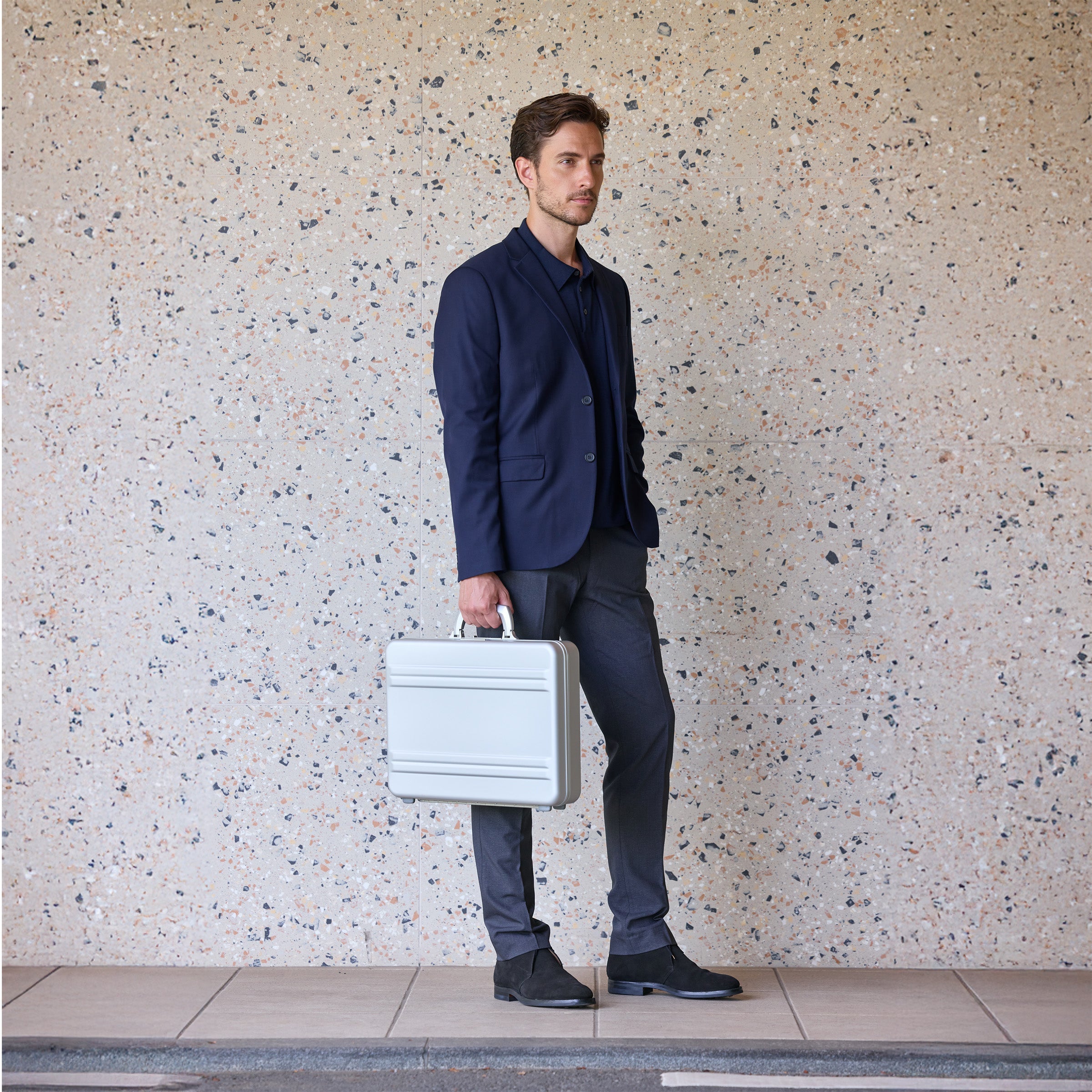 Man in a navy suit holding a silver briefcase against a speckled wall.