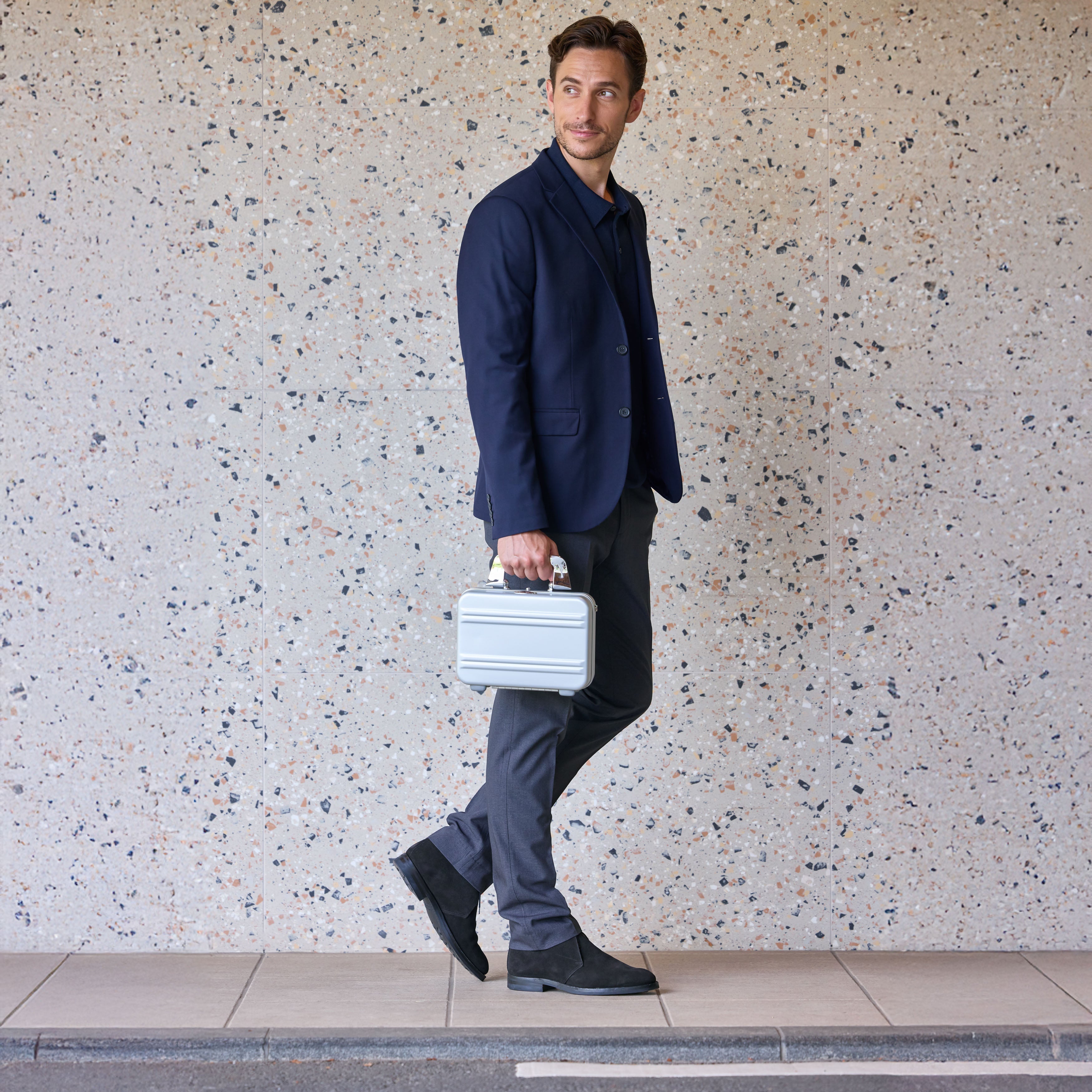 Man in a navy suit holding a white briefcase against a textured wall.