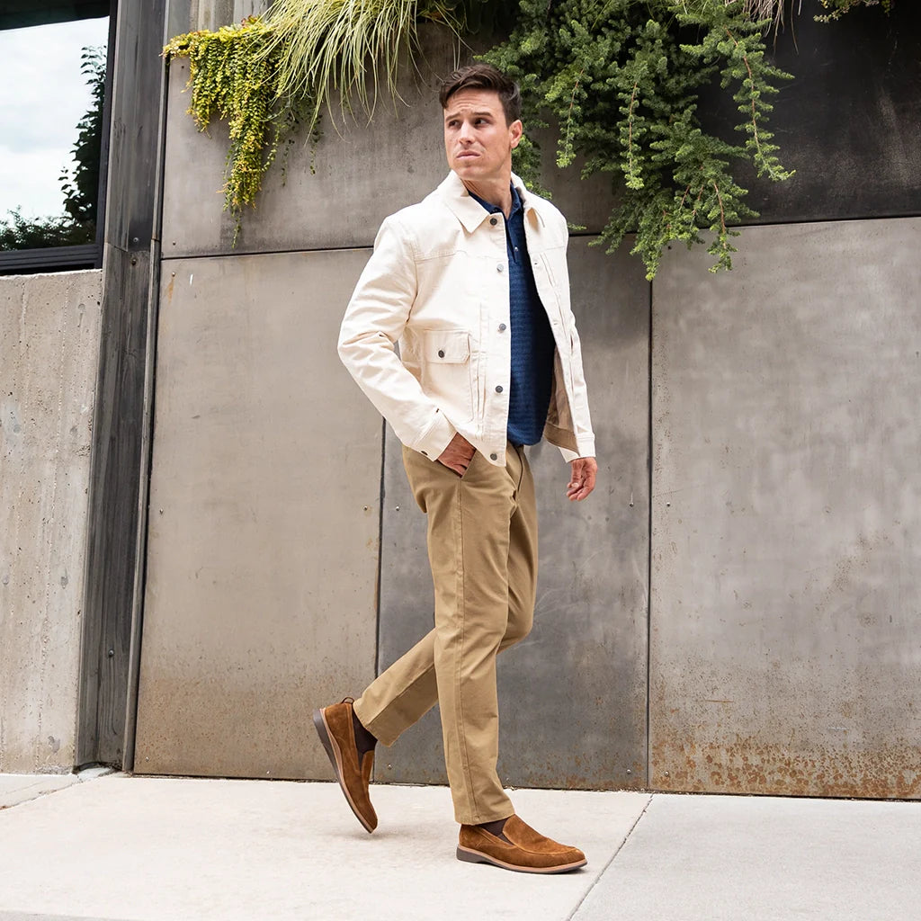 Man in a white jacket and beige pants walking outdoors with plants in the background