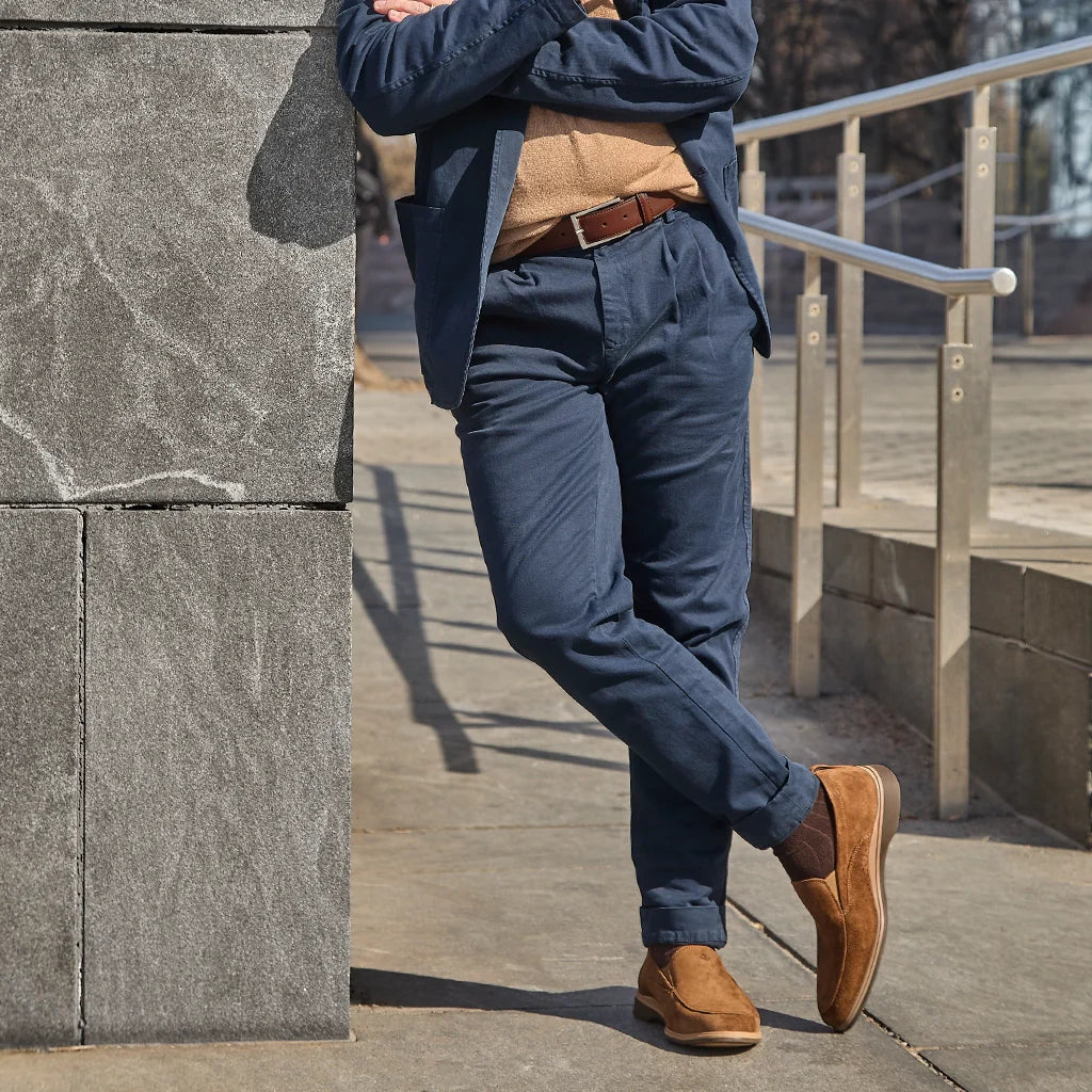 Person wearing a blue blazer, beige sweater, blue pants, and brown shoes leaning against a stone wall.