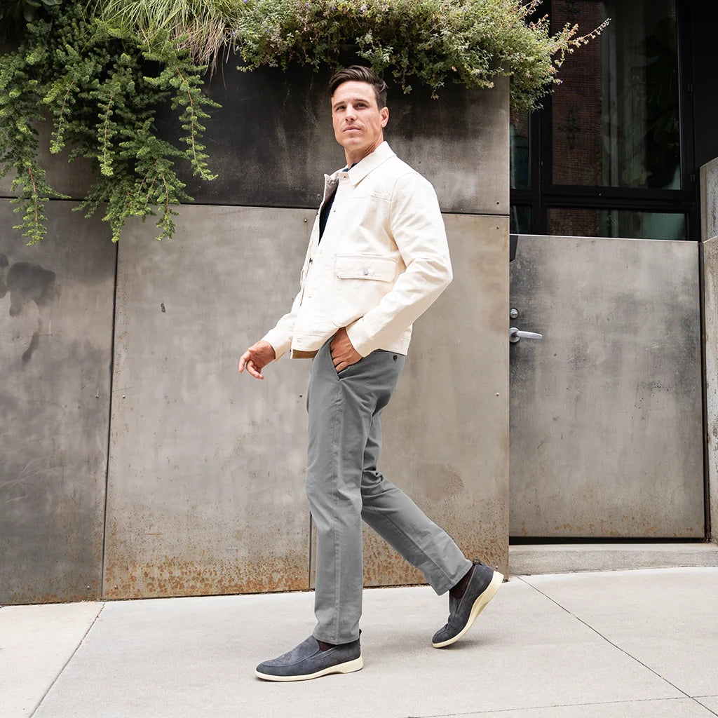 Man in a white jacket and gray pants walking outdoors against a concrete wall with plants.