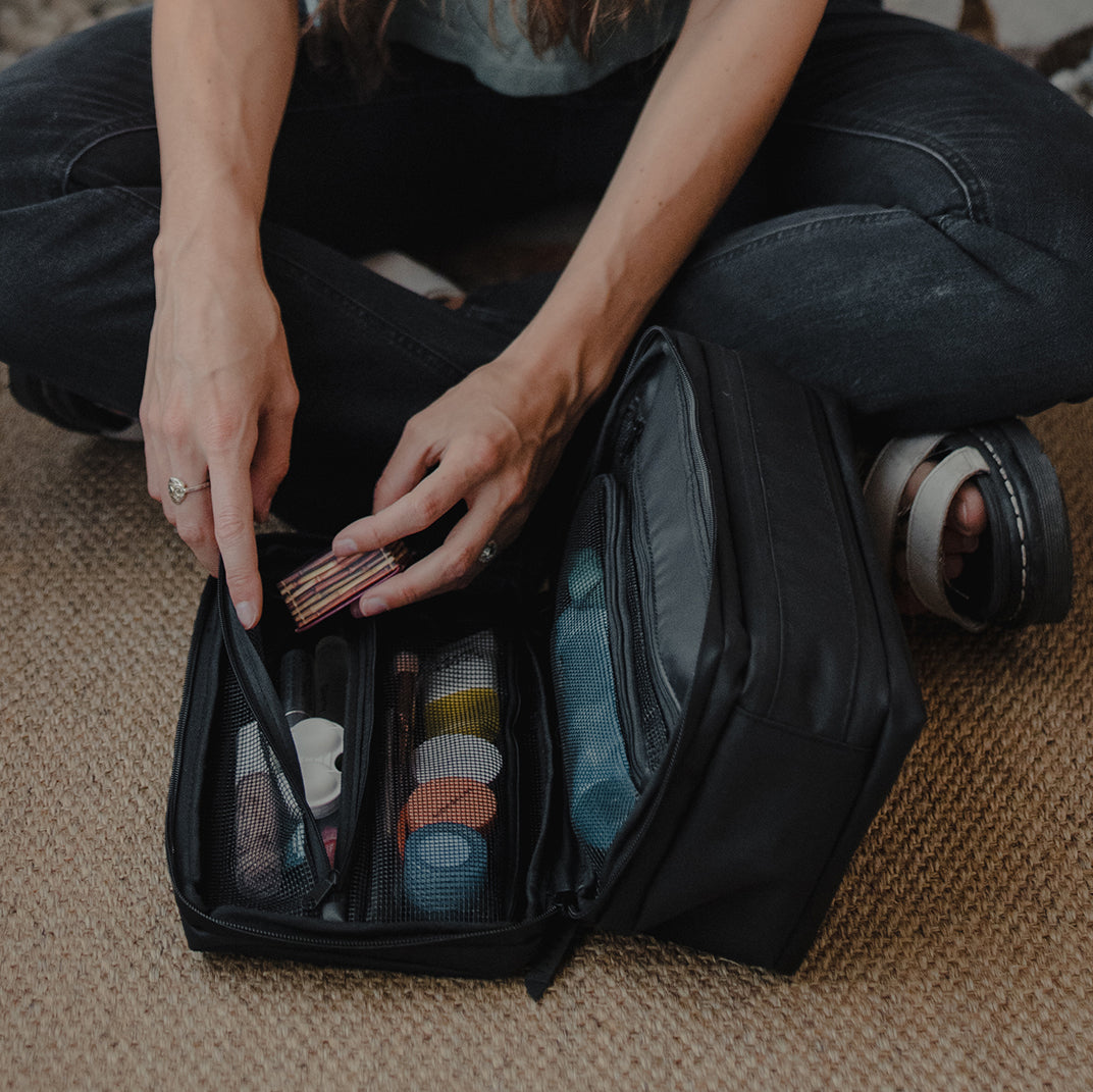 Person organizing makeup brushes in a black travel bag on a carpeted floor.