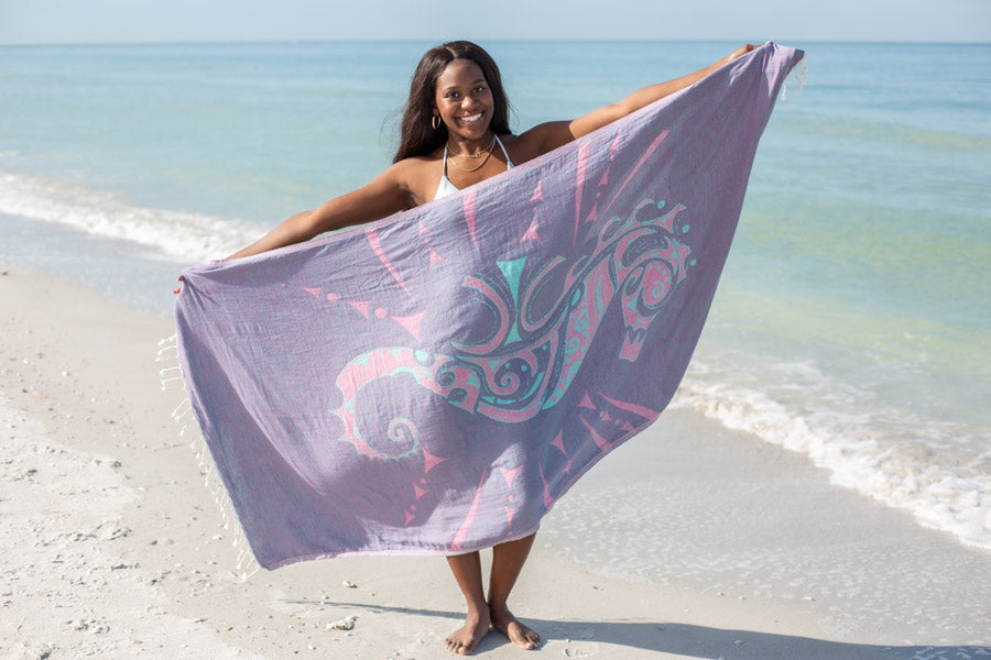 Woman holding a beach towel with a seahorse design on a sandy beach.