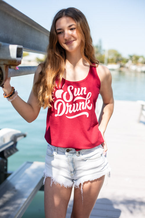 Woman wearing a red tank top with 'Sun Drunk' text by a waterfront.