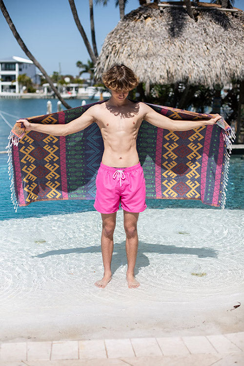 Person in pink swim shorts standing with a colorful towel draped over shoulders by a pool.
