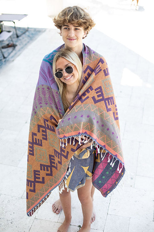 Two people wrapped in a colorful patterned blanket on a white floor.