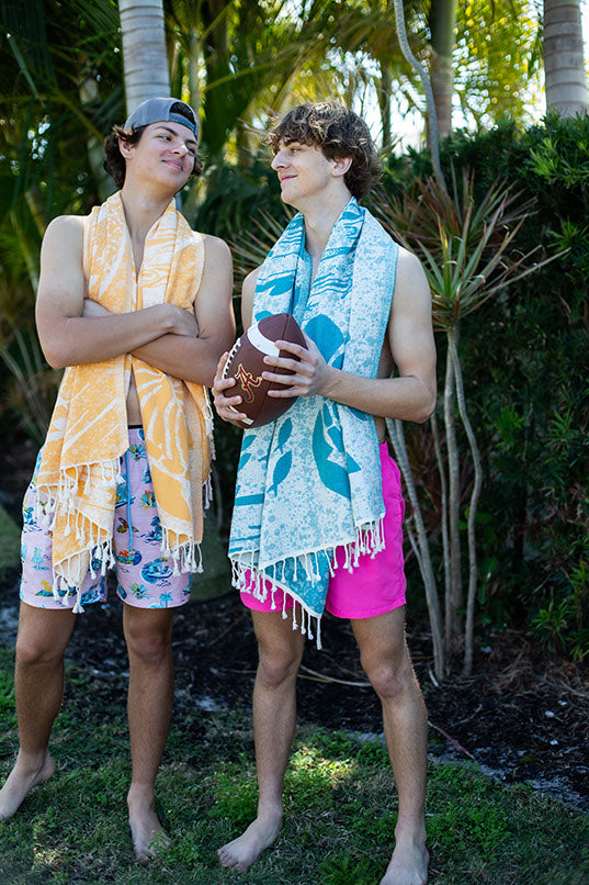 Two people standing outdoors with towels draped over their shoulders, surrounded by greenery.