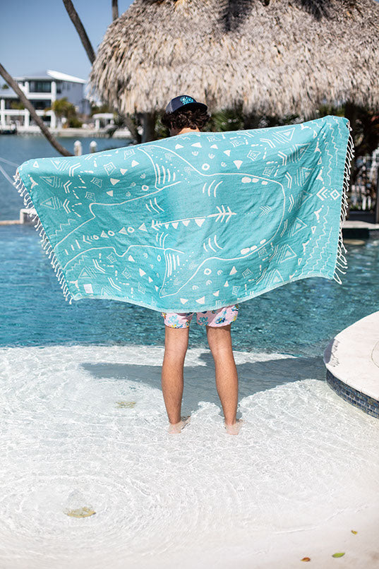 Person holding a turquoise towel with white patterns by a poolside.