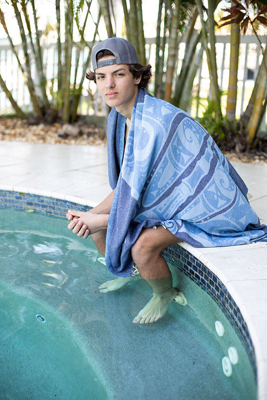 Person wrapped in a blue towel sitting by a pool with plants in the background