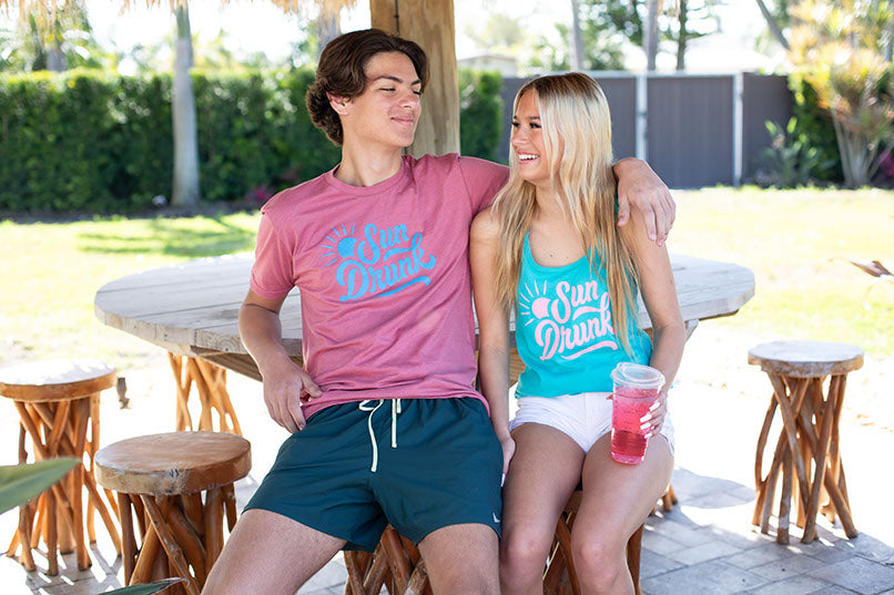 Two people sitting outdoors with 'Sun Drawl' shirts and drinks.