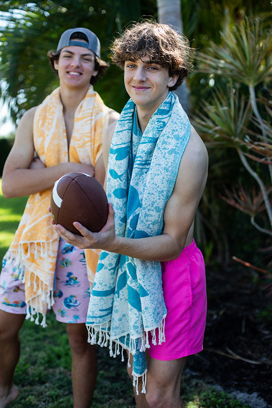 Two people standing outdoors with towels and a football, surrounded by greenery.